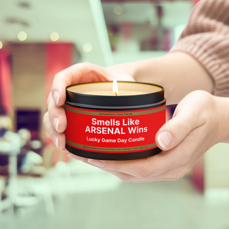 Wonderscape Wicks candle shop on Etsy presenting lit 8 oz tin candle with text smells like Arsenal Wins Lucky Game Day Candle held by a woman on a lifestyle background. The all natural cotton-wick candle can be personalized with customer's custom text. The design is aimed at Premier League Football Soccer fans with Arsenal red and white colors. Gift for wife boyfriend husband girlfriend brother sister Bukayo Saka Martin Odegaard Declan Rice Gabriel Martinelli Thierry Henry Dennis Bergkamp