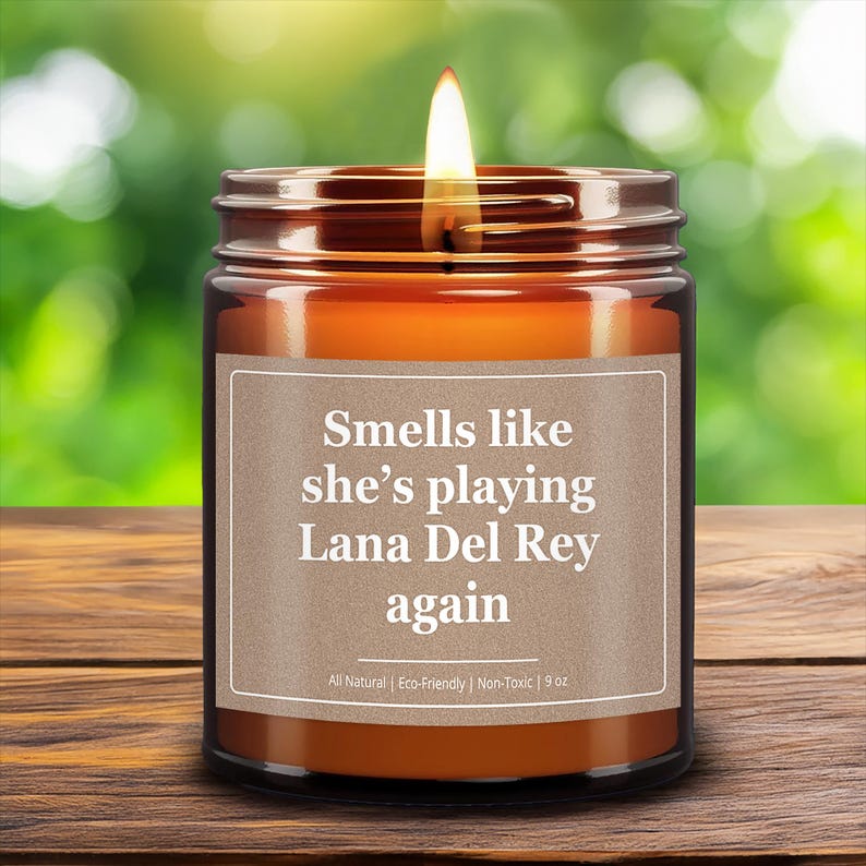 Wonderscape Wicks candle shop on Etsy presenting 9 oz amber jar soy wax lit candle with text smells like she is playing Lana Del Rey again on a wood table in a garden. LDR music fan gift for wife girlfriend mom bestie Billie Eilish Taylor Swift