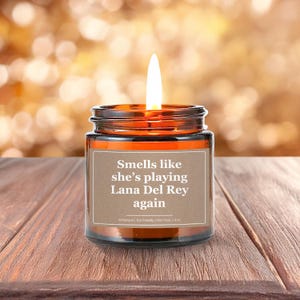 Wonderscape Wicks candle shop on Etsy presenting 4 oz amber jar soy wax lit candle with text smells like she is playing Lana Del Rey again on a wood table in a garden. LDR music fan gift for wife girlfriend mom bestie boss Ed Sheeran Eminem Madonna