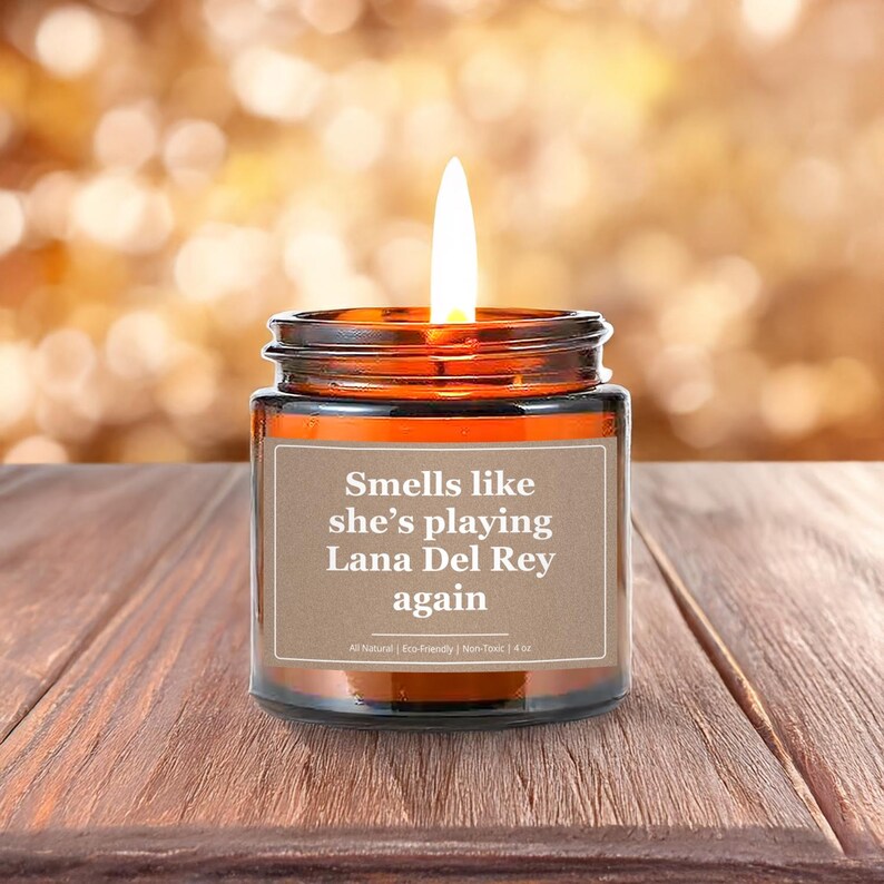 Wonderscape Wicks candle shop on Etsy presenting 4 oz amber jar soy wax lit candle with text smells like she is playing Lana Del Rey again on a wood table in a garden. LDR music fan gift for wife girlfriend mom bestie boss Ed Sheeran Eminem Madonna