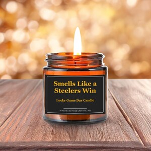 Wonderscape Wicks candle shop on Etsy presenting lit 4 oz amber jar soy wax candle with text smells like a Steelers win lucky game day candle on a wood table in a garden. Pittsburgh Football NFL sports gift for husband wife fiance boyfriend