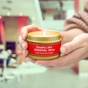 Wonderscape Wicks candle shop on Etsy presenting lit 4 oz tin candle with text smells like Arsenal Wins Lucky Game Day Candle held by a woman on a lifestyle background. The all natural cotton-wick candle can be personalized with customer's custom text. The design is aimed at Premier League Football Soccer fans with Arsenal red and white colors. Gift for wife boyfriend husband girlfriend brother sister Bukayo Saka Martin Odegaard Declan Rice Gabriel Martinelli Thierry Henry Dennis Bergkamp