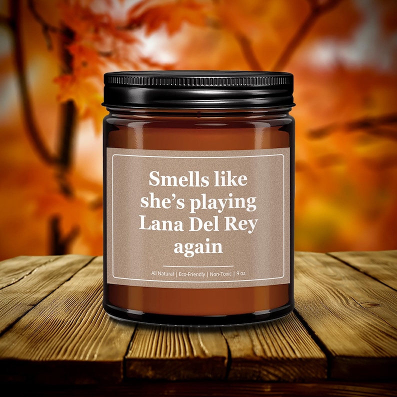 Wonderscape Wicks candle shop on Etsy presenting black lid 9 oz amber jar soy wax lit candle with text smells like she is playing Lana Del Rey again on a wood table in a garden. LDR music fan gift for wife girlfriend mom bestie boss Beyonce Dua Lipa