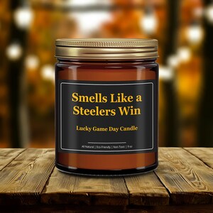 Wonderscape Wicks candle shop on Etsy presenting gold lid 9oz amber jar soy wax candle with text smells like a Steelers win lucky game day candle on a wood table in a garden. Pittsburgh Football NFL sports gift for husband wife fiance boyfriend