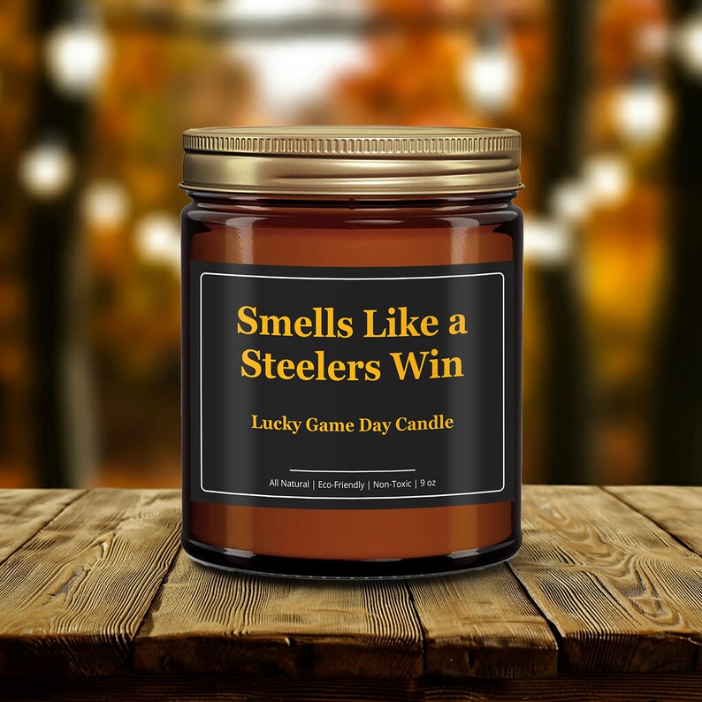 Wonderscape Wicks candle shop on Etsy presenting gold lid 9oz amber jar soy wax candle with text smells like a Steelers win lucky game day candle on a wood table in a garden. Pittsburgh Football NFL sports gift for husband wife fiance boyfriend