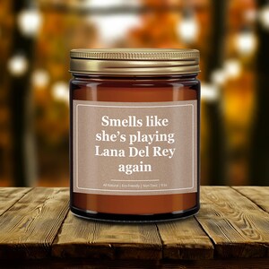 Wonderscape Wicks candle shop on Etsy presenting gold lid 9 oz amber jar soy wax lit candle with text smells like she is playing Lana Del Rey again on a wood table in a garden. LDR music fan gift for wife girlfriend mom bestie Ariana Grande Weeknd