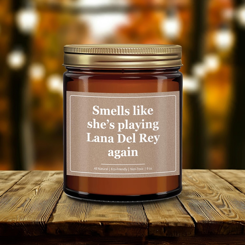 Wonderscape Wicks candle shop on Etsy presenting gold lid 9 oz amber jar soy wax lit candle with text smells like she is playing Lana Del Rey again on a wood table in a garden. LDR music fan gift for wife girlfriend mom bestie Ariana Grande Weeknd