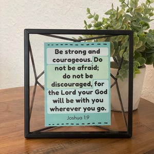 Puede incluir: Un letrero enmarcado en metal negro con el texto "S&eacute; fuerte y valiente. No temas; no te desanimes, porque el Se&ntilde;or tu Dios estar&aacute; contigo dondequiera que vayas. Josu&eacute; 1:9" sobre un fondo azul claro y verde.
