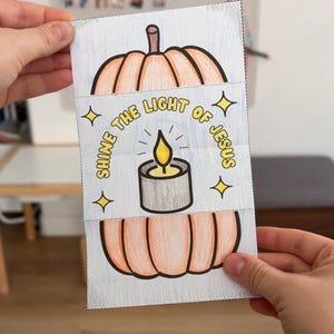 May include: A rectangular paper craft featuring a hand-coloured orange pumpkin divided into two sections. The centre section displays a grey candle with a yellow flame and the text SHINE THE LIGHT OF JESUS in yellow, surrounded by four yellow stars.