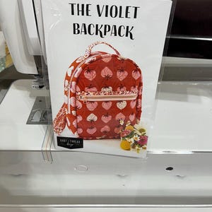 May include: A sewing pattern titled "THE VIOLET BACKPACK" is displayed. The backpack design features a red background with a pattern of pink strawberries. It has a pink zipper and a small handle. A small bouquet of flowers is placed near the bottom.