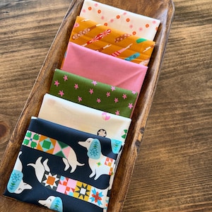 May include: A stack of six folded fabric squares featuring various patterns including a white background with small orange flowers, a mustard yellow print, a solid pink, a dark green with pink stars, a cream print, and a black dachshund dog print.