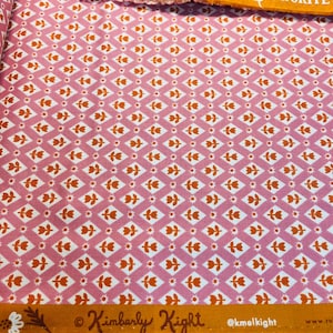 May include: Pink fabric with a repeating pattern of orange and white stylized flowers. The fabric has a gold label with the text "RUBY STAR SOCIETY" and "FAVORITE" in white. The bottom of the fabric has a gold label with the text "Kimberly Kight" and "@kmelkight".