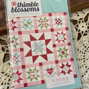 May include: A quilt pattern for a 84-inch star quilt called "ADORE" by Thimble Blossoms, designed by Camille Roskelley. The pattern features a red, pink, and green color scheme with a variety of star and heart blocks.