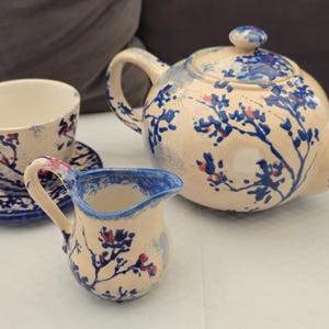 Peut inclure: Service à thé en céramique crème avec des motifs floraux bleus et des touches de rouge. L'ensemble comprend une théière, un pot à crème, une tasse et une soucoupe. La théière est arrondie et possède un couvercle.