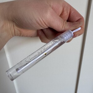 Peut inclure: Un flacon cylindrique transparent contenant un liquide, tenu dans une main. Le flacon a un bouchon blanc et un applicateur blanc. Le texte "Serene & Cherished" est imprimé sur le flacon en écriture cursive.