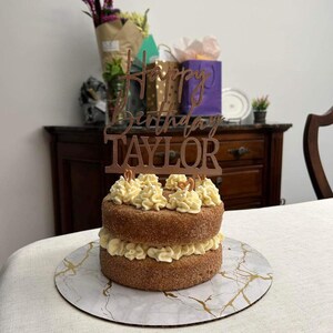 Könnte beinhalten: Ein zweistöckiger Kuchen mit Vanille-Frosting und einer Zimt-Zucker-Beschichtung. Der Kuchen ist mit einem goldenen Topper verziert, auf dem "Happy Birthday Taylor" steht.
