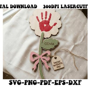 Puede incluir: Artesanía de flores de madera con una huella de mano roja, una hoja verde con el nombre "Olivia" y un lazo rosa. Una etiqueta con "happy mother's day" cuelga de la flor. Las palabras "DIGITAL DOWNLOAD" y "300DPI LASERCUTFILE" están en la parte superior.