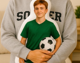 May include: A white, body-shaped pillow with a printed image of a young person wearing a green V-neck shirt and holding a soccer ball. The person is embraced by someone wearing a gray sweatshirt with the word "SOCCER" printed on it.