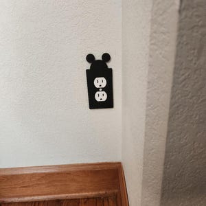 May include: Black outlet cover with a Mickey Mouse silhouette design. The cover is mounted on a white wall, with a wooden door frame visible at the bottom. The outlet itself has two standard American plugs.