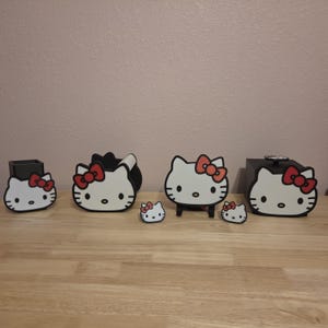 May include: A set of black and white desk accessories featuring the Hello Kitty character. The collection includes a pen holder, a tape dispenser, two small decorative accents, a small display stand, and a square box, all with red bows.