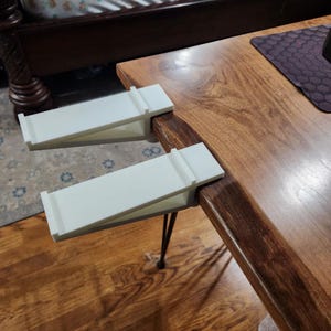May include: Two white, angled table clamps are attached to the edge of a wooden table. The clamps are designed to hold items in place. The table has a natural wood grain finish and is supported by black metal legs.