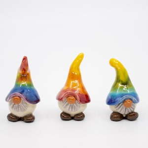 Può includere: Tre gnomi in ceramica con cappelli arcobaleno. I gnomi sono in piedi su una superficie bianca.