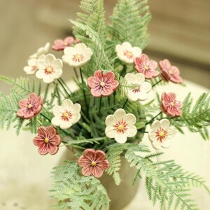 Peut inclure: Un bouquet de fleurs en céramique dans des tons de rose et de blanc, disposé dans une verdure ressemblant à une fougère. Les fleurs ont un aspect délicat et vintage.