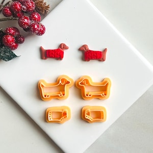 Puede incluir: Dos cortadores de galletas de dachshund rojos y dos cortadores de galletas de dachshund naranjas sobre una superficie blanca.