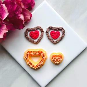 May include: Four heart-shaped cookie cutters, two brown with red hearts inside and two orange with no hearts inside. The cutters are on a white surface.