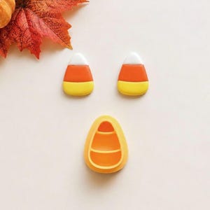 Peut inclure: Deux petites breloques triangulaires en forme de bonbon candy corn avec des bandes horizontales blanches, orange et jaunes, accompagnées d'un moule ou emporte-pièce jaune, plus grand, creux et triangulaire avec des crêtes internes.