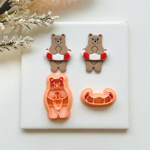 May include: A selection of bear-shaped cookie cutters and bear-themed decorations. Two brown bears wear red and white life preservers. There is a bear-shaped cookie cutter and a life preserver-shaped cutter. The items are on a white surface.
