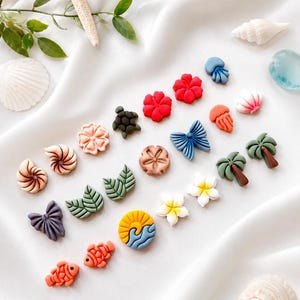 May include: A collection of small, colourful stud earrings featuring various shapes including red hibiscus flowers, blue butterflies, green palm trees, orange fish, sea shells, leaves, a sun and wave design, and jellyfish on a white fabric background.