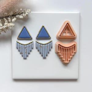 May include: Earrings and cutters in a geometric design. The earrings have a blue triangle top with a gold border, and a light blue, fan-shaped bottom with gold accents. Two peach-coloured cutters are also shown, one triangle and one fan-shaped.