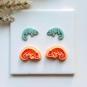 May include: Four chameleon-shaped items. Two are light teal-colored chameleons. Below are two orange-colored chameleon cookie cutters. The items are arranged on a white surface, with a sprig of dried flowers in the upper left corner.
