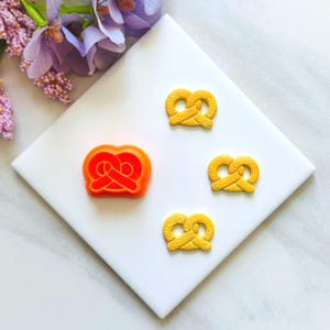 Puede incluir: Un cortador de galletas naranja con forma de pretzel junto a tres galletas amarillas con forma de pretzel. Las galletas están sobre una superficie blanca con flores moradas en el fondo. El cortador es de plástico.