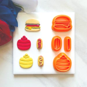 Op de afbeelding: Een assortiment voedselthema koekjessnijders en koekjes. De afbeelding toont een afgewerkte hamburger en hotdogs, samen met oranje plastic snijders voor een hamburger, friet en een pompoenvormig koekje. De koekjes zijn rood, geel en bruin.