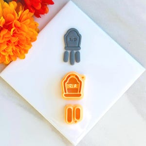 May include: Two sets of Halloween cookie cutters. One set is gray and features a tombstone with the text "RIP" and three small rectangular shapes. The other set is orange and features a tombstone with the text "RIP" and two small rectangular shapes.