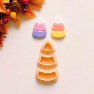 Könnte beinhalten: Zwei Ornamente in Form von Zuckermais und drei gestapelte Ausstechformen aus orangefarbenem Kunststoff. Ein Ornament ist weiß, rosa und lila, das andere weiß, orange und gelb. Die drei orangefarbenen Ausstechformen sind dreieckig und gebogen.
