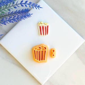 Può includere: Tre contenitori di popcorn, uno rosso e bianco con popcorn bianco, e due arancioni con popcorn giallo. I contenitori sono in resina e sono perfetti per aggiungere un tocco divertente a qualsiasi progetto artigianale.
