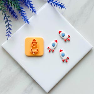 May include: An orange rocket-shaped cookie cutter and three white, red, and blue rocket-shaped decorations are displayed on a white surface. The cookie cutter is square. The rockets have red fins and nose cones.