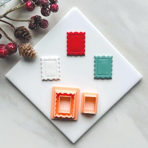 May include: A collection of colorful, stamp-shaped cookie cutters in red, white, teal, and peach. The cutters are arranged on a white surface, with a sprig of berries in the upper left corner. The cutters have a scalloped edge.