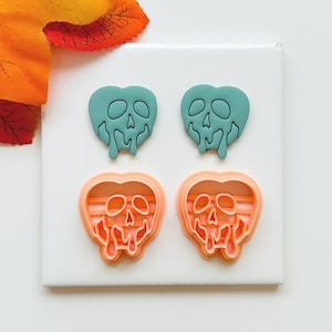 May include: Four heart-shaped cookie cutters. Two are teal, and two are orange. Each cutter features a skull design with dripping details. The cutters are arranged on a white surface, with orange and yellow leaves in the background.