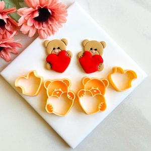 Puede incluir: Dos cortadores de galletas de osito de peluche marrón que sostienen formas de corazón rojo, y dos cortadores de galletas en forma de corazón. Todos están sobre una superficie blanca.