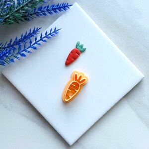 Puede incluir: Dos amuletos de arcilla polimérica en forma de zanahoria naranja y roja. Los amuletos están sobre un fondo blanco.