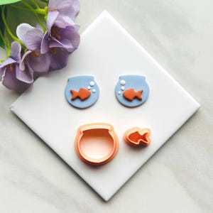 Puede incluir: Un conjunto de cortadores de galletas y decoraciones con temática de peces en naranja y azul. El conjunto incluye dos peceras azules con peces y burbujas naranjas, además de un cortador con forma de pecera y pez. Flores moradas en el fondo.
