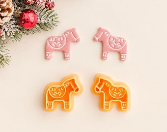 May include: Two pink horse-shaped cookie cutters and two orange horse-shaped cookie cutters. Each cutter features a detailed saddle design with intricate patterns on the back. The items are arranged in two rows on a plain surface near pine needles.