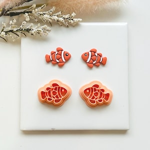 May include: Four orange and white clownfish-shaped items on a white surface. Two are solid, and two are layered. The clownfish have black eyes and white stripes. The background includes white flowers and dried grass.