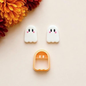 May include: Two white ghost-shaped clay pieces with black oval eyes and pink circular cheeks, positioned above a single light orange plastic ghost-shaped cookie cutter with a hollow center and a small horizontal bar across the top.