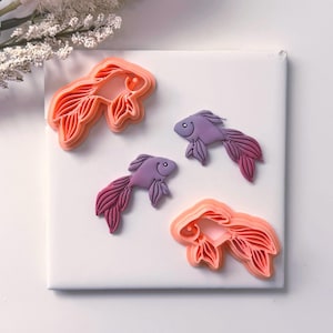 May include: Four fish-shaped cookie cutters in shades of orange and purple. Two cutters are orange, and two are purple. The fish have detailed fins and tails. The cutters are arranged on a white surface.