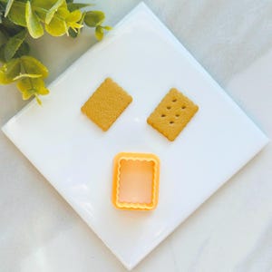 May include: Three cookie cutters on a white surface. Two are shaped like rectangles and one is shaped like a square. All are a light orange color.